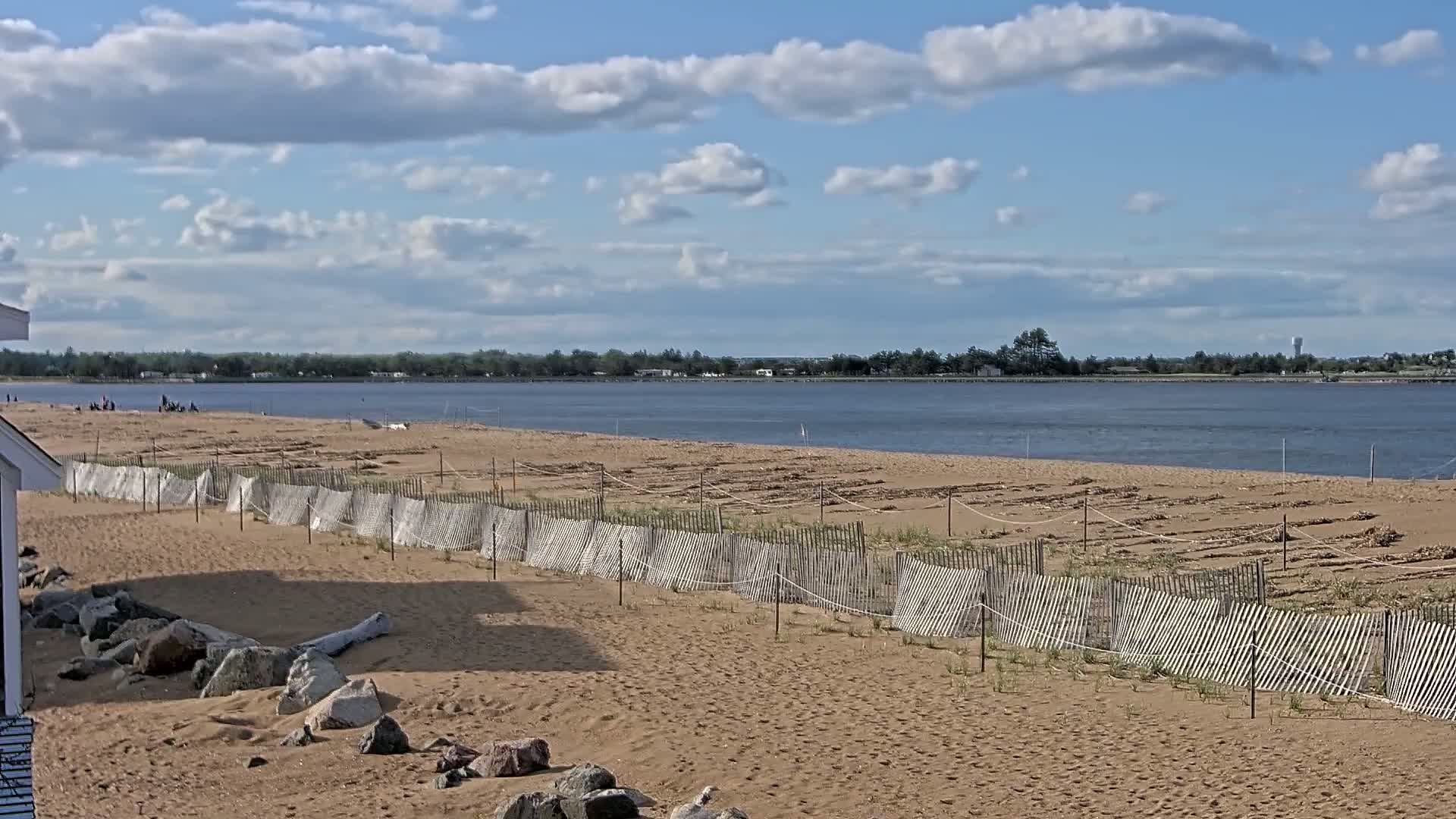 A sandy beach with a low wooden fence separating it from a calm body of water under a partly cloudy sky.
