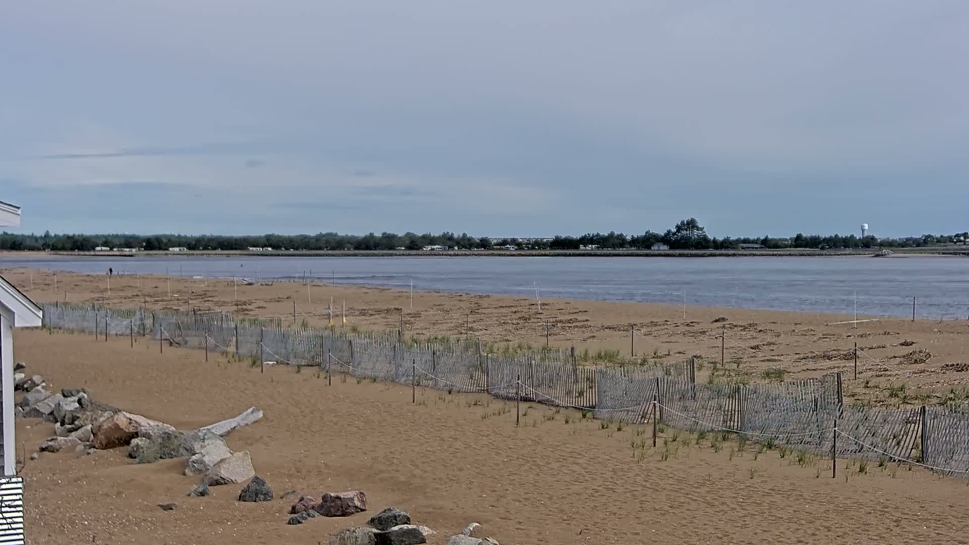 A sandy beach with a low wooden fence and rocks stretches to a calm body of water under a mostly cloudy sky.
