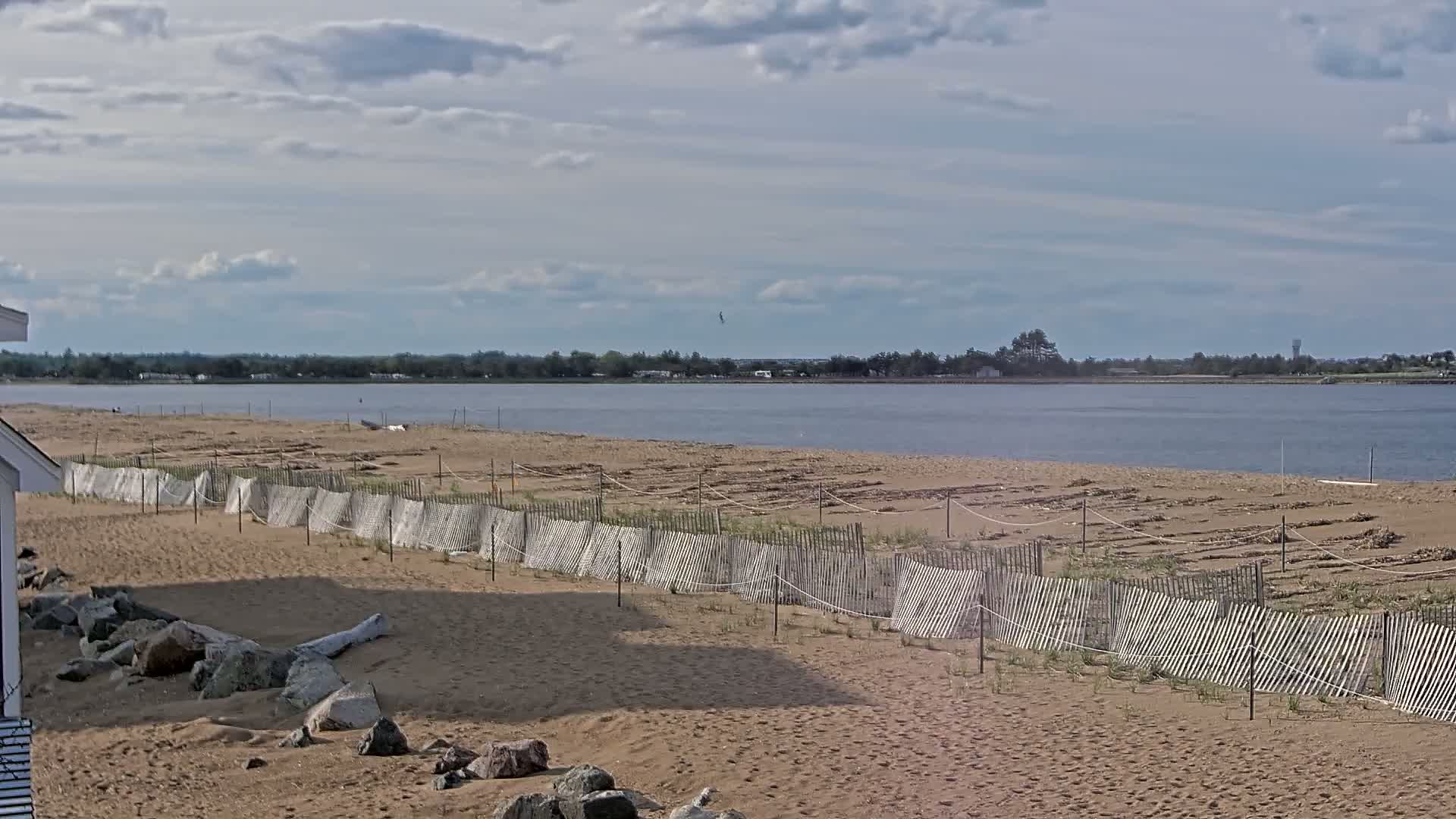 A sandy beach with a low wooden fence stretches along a calm body of water under a partly cloudy sky.