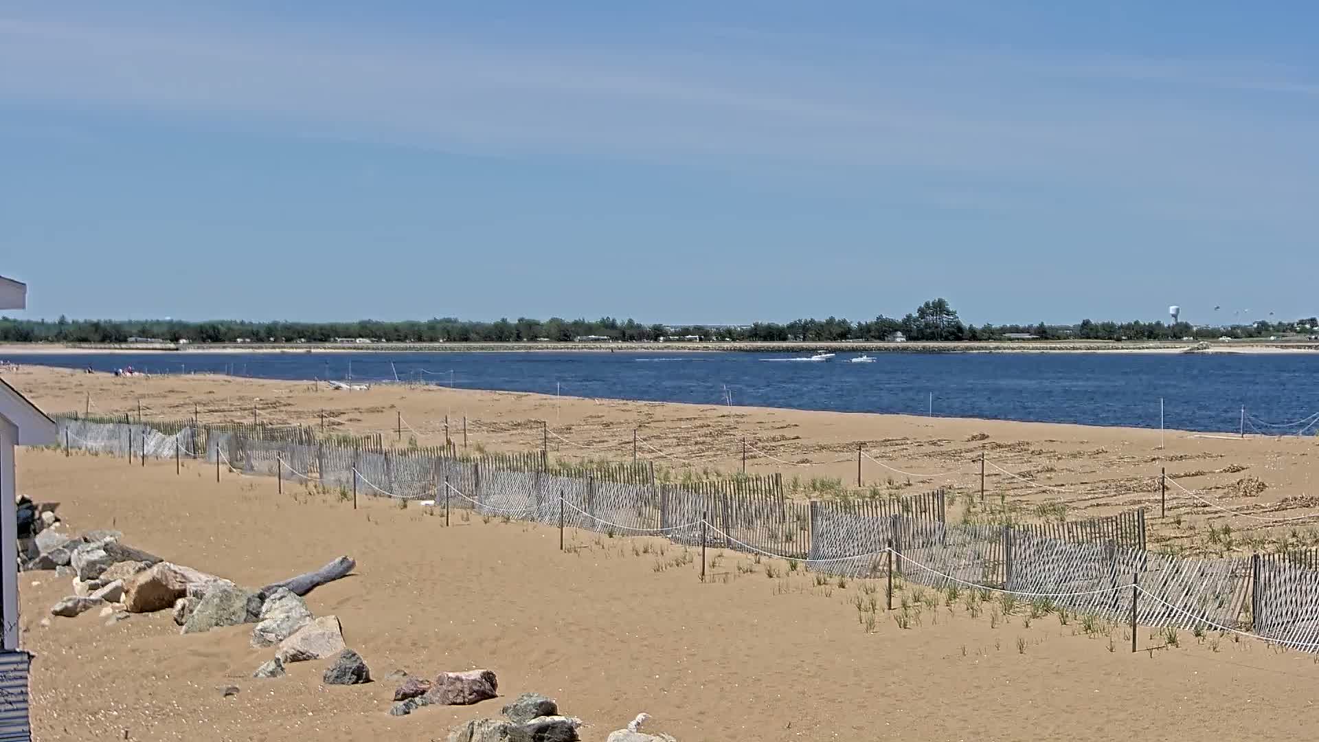 A sandy beach with a wooden fence and rocks stretches toward a calm body of water under a clear blue sky.