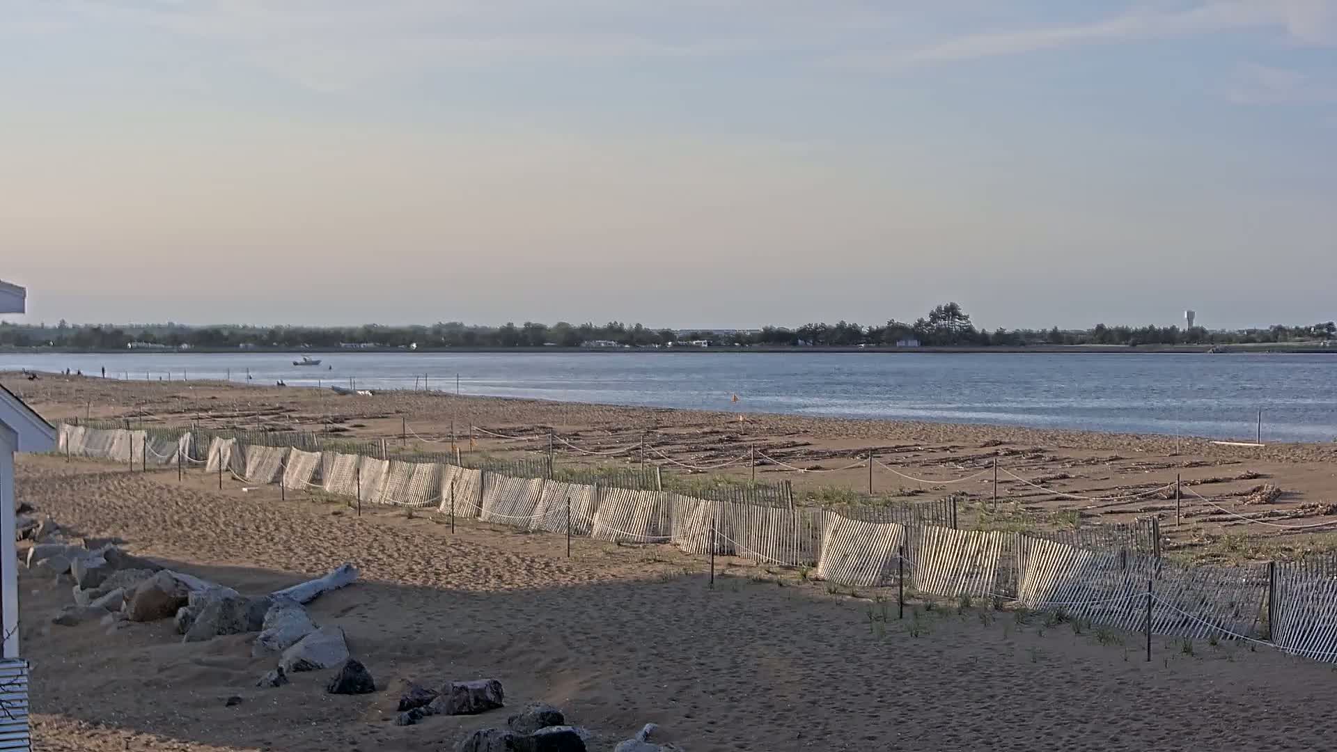 A sandy beach with a wooden fence, seaweed strewn across the sand, and a calm body of water in the background under a mostly clear sky.