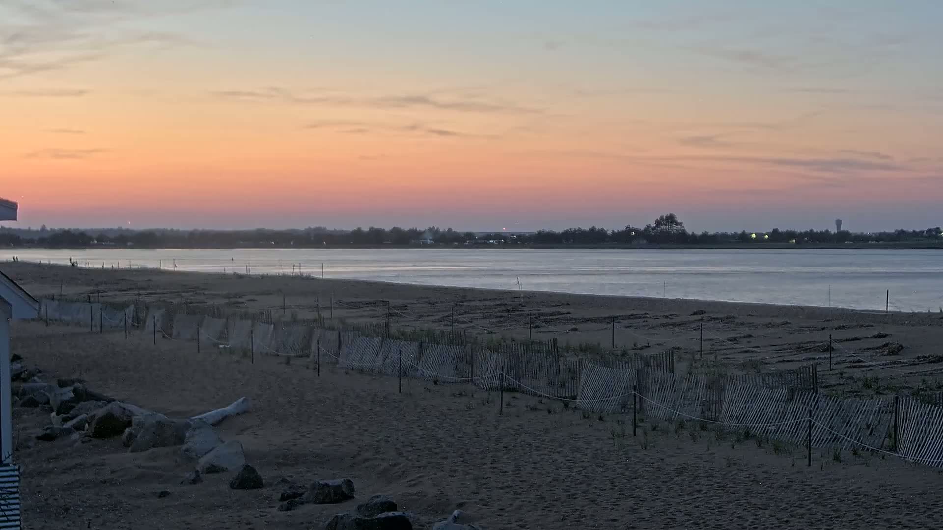 A sandy beach with short fences stretches along a calm body of water under a pink and orange sunset.