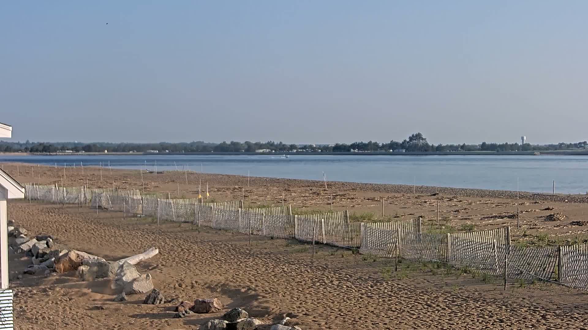 A sandy beach with low wooden fences stretches toward a calm body of water under a clear, bright sky.
