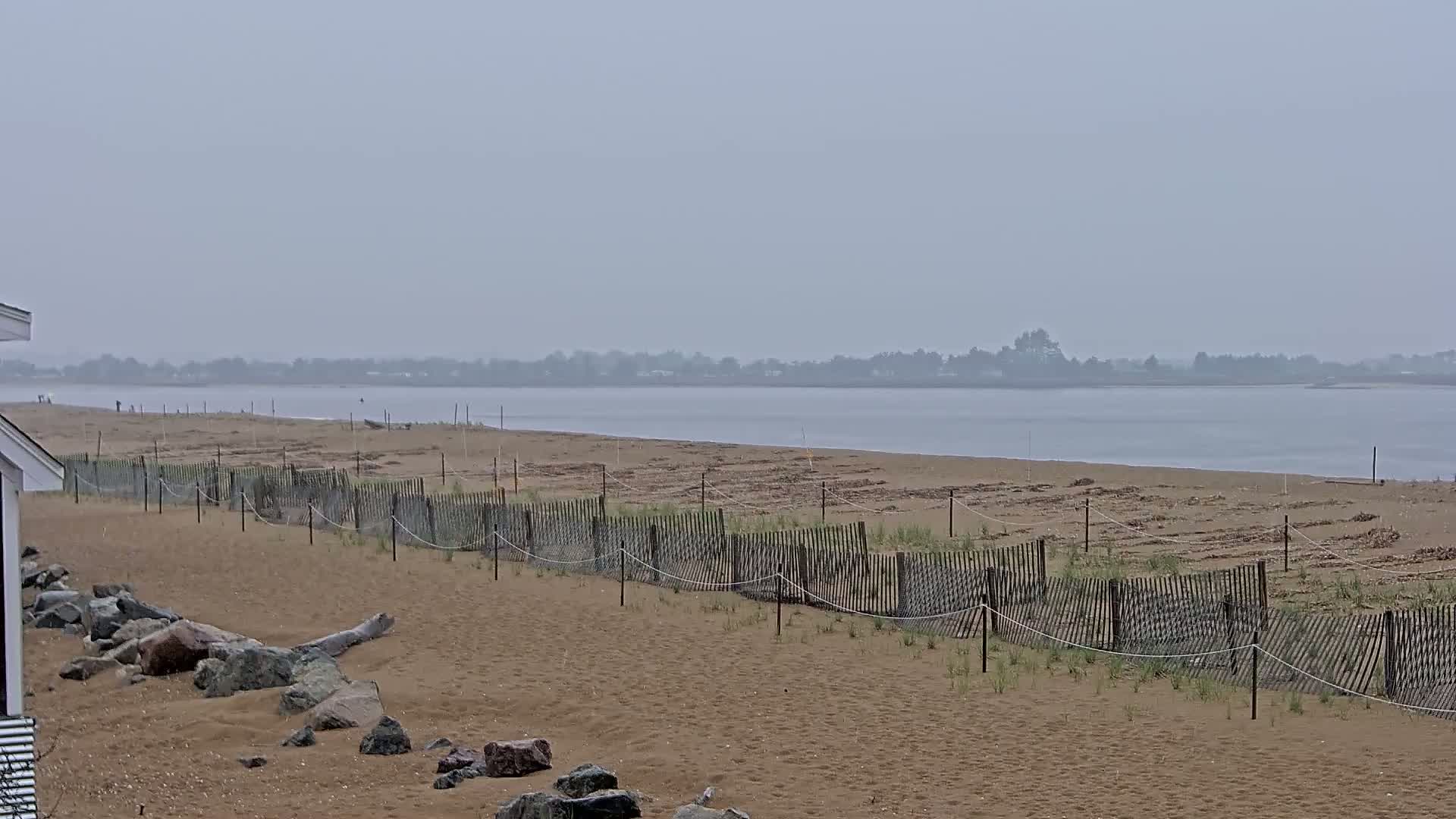 A sandy beach with a short wooden fence stretches along a calm body of water under an overcast sky.
