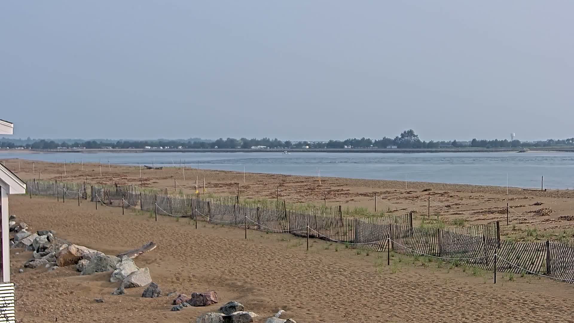 A sandy beach with a wooden fence stretches along a calm, light-blue body of water under a hazy sky.