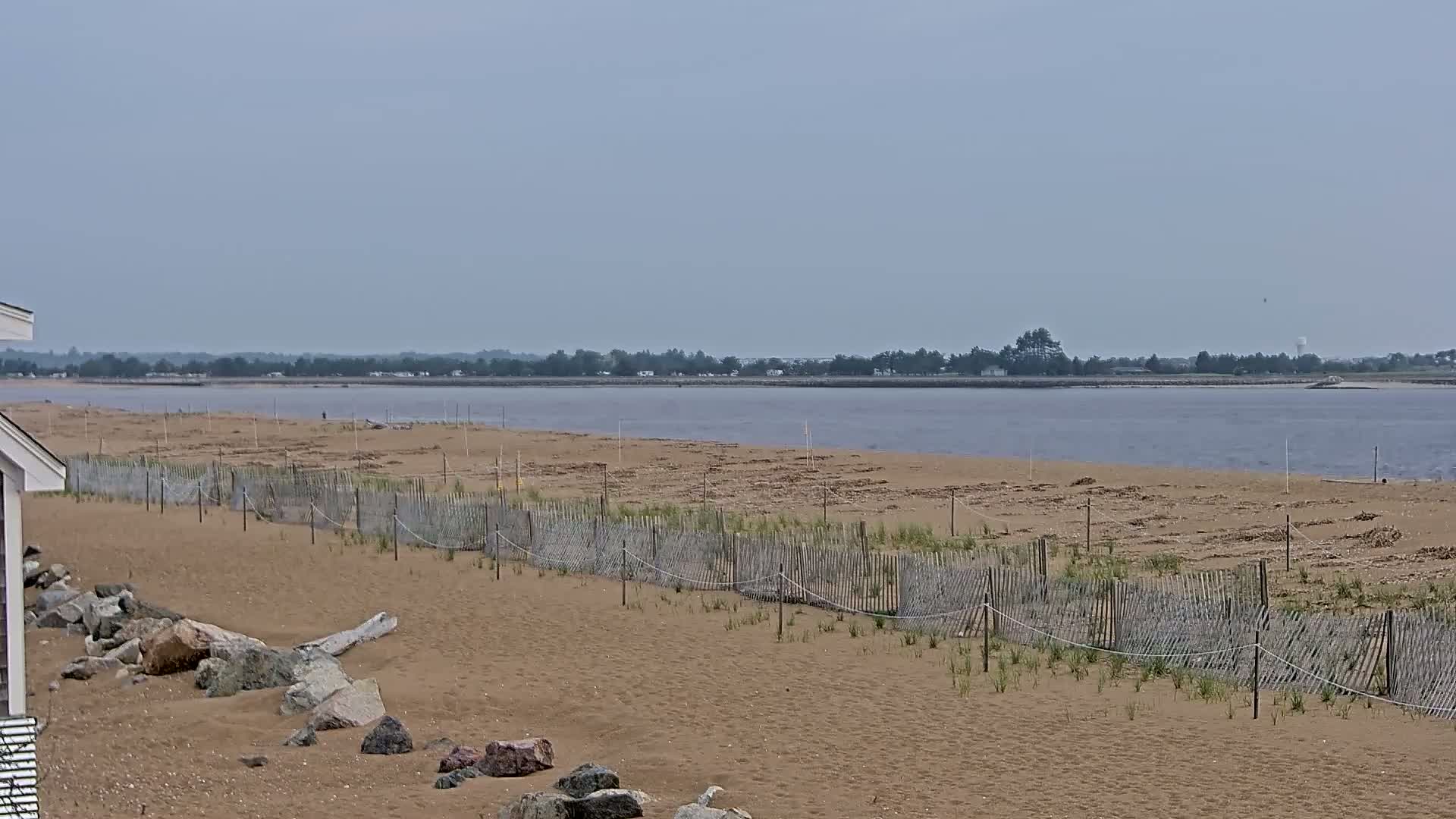 A sandy beach with a low wooden fence stretches toward a calm, wide body of water under a cloudy sky.