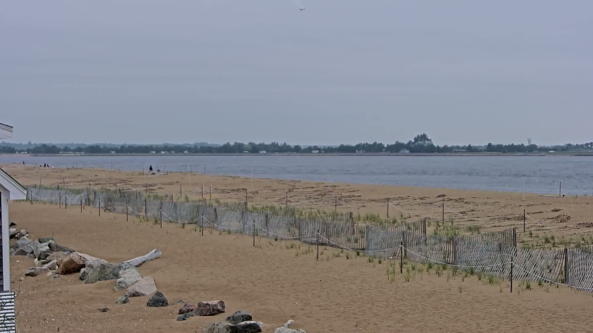 A sandy beach with a low wooden fence stretches toward a calm body of water under an overcast sky.