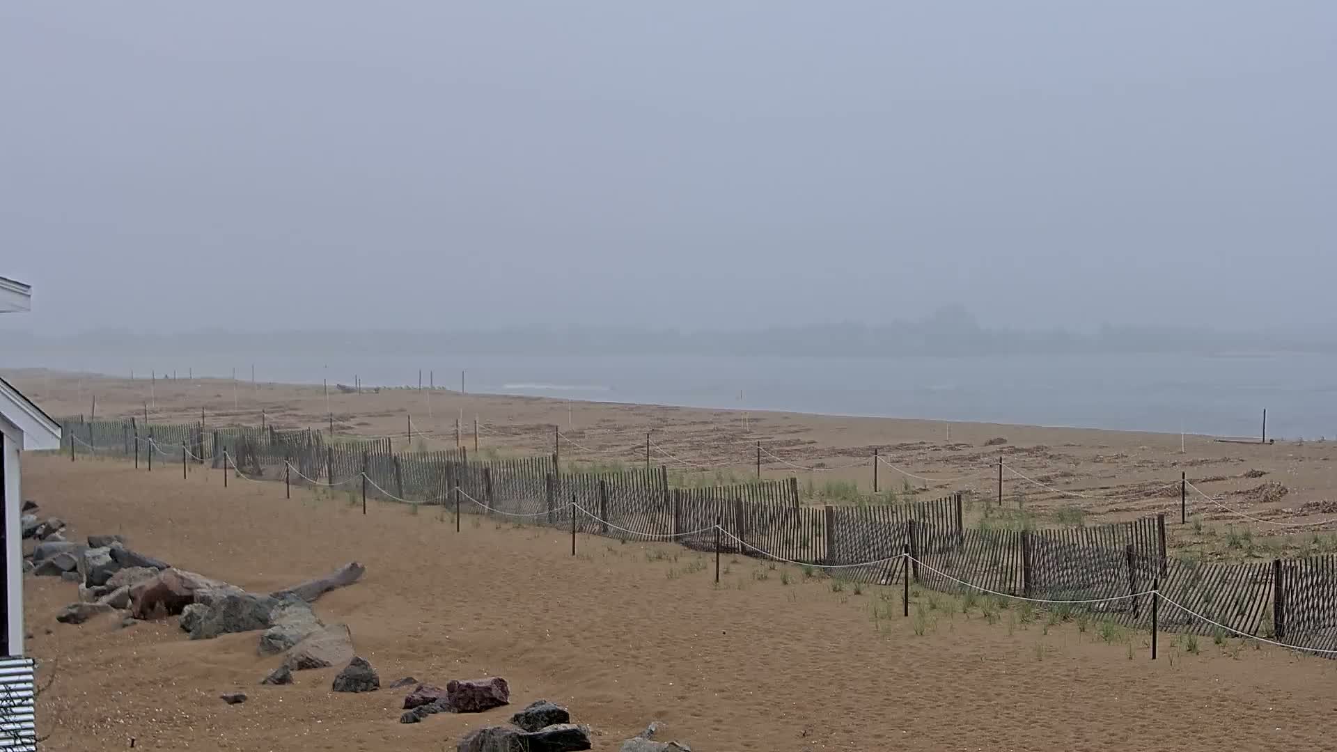 A foggy beach with a wooden fence and scattered rocks is seen from a slightly elevated vantage point.