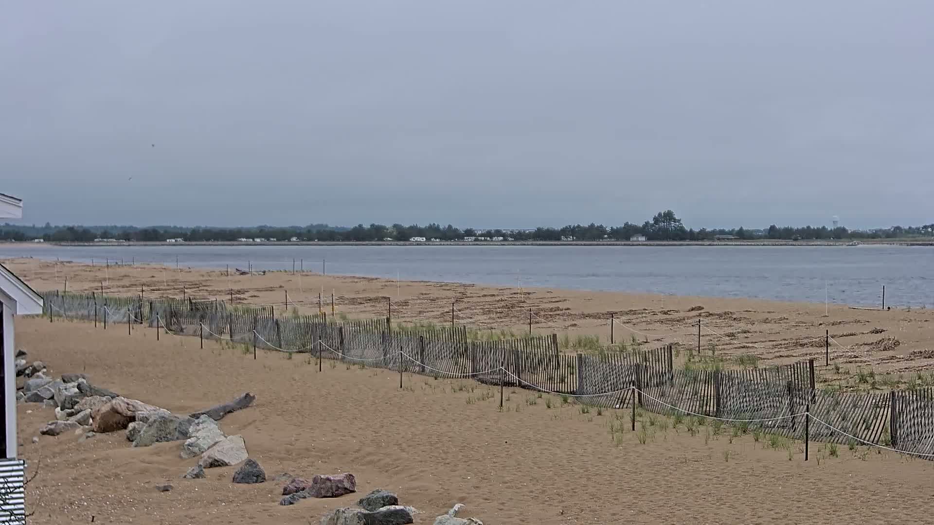A sandy beach with a low wooden fence stretches along a calm body of water under an overcast sky.