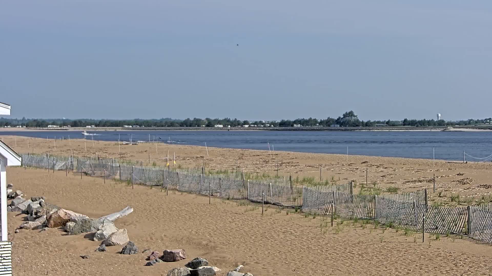 A sandy beach with a low wooden fence stretches along a calm body of water under a clear sky.