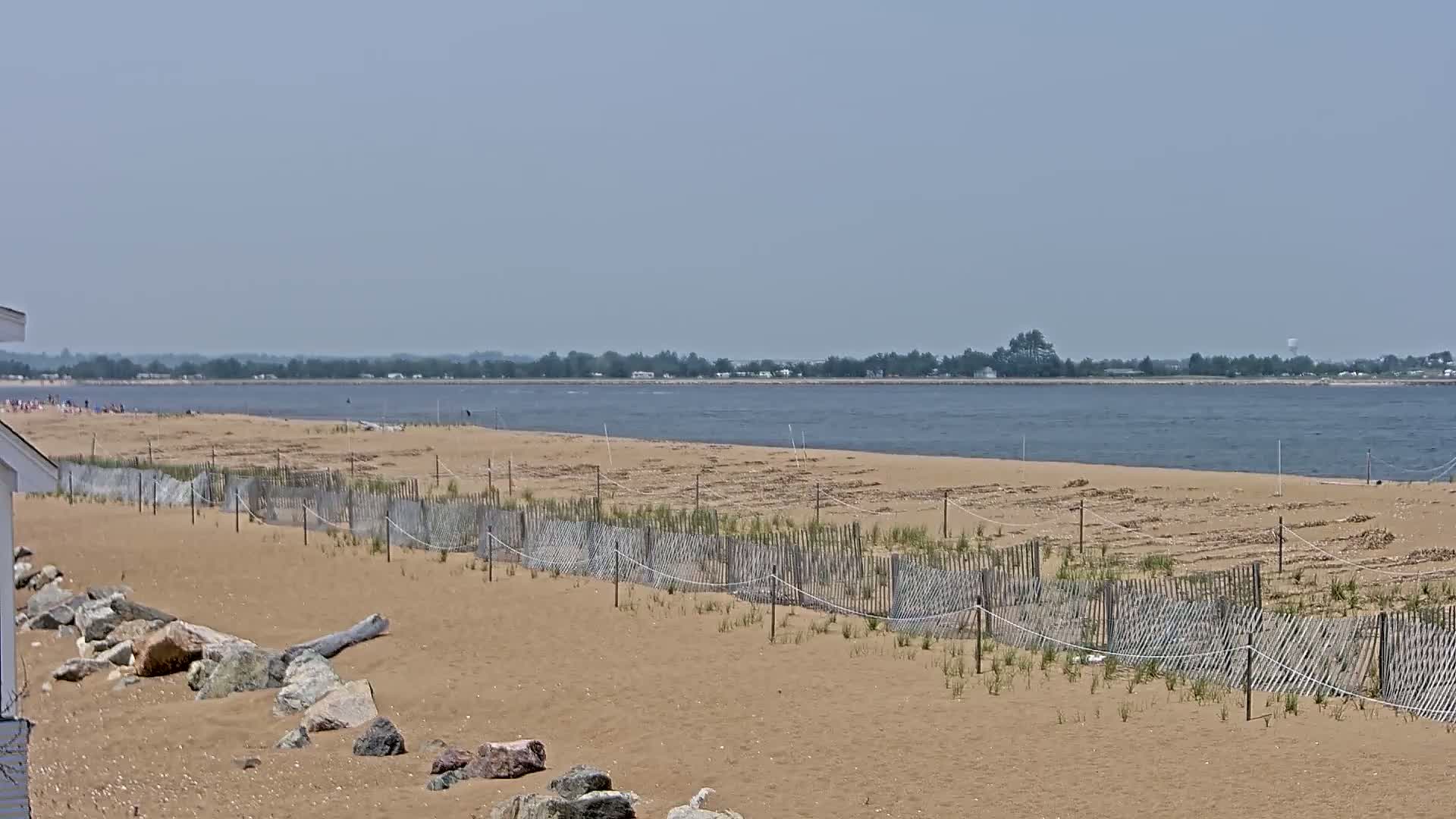 A sandy beach with a wooden fence stretches along a calm body of water under a hazy sky, with a distant tree line visible on the opposite shore.