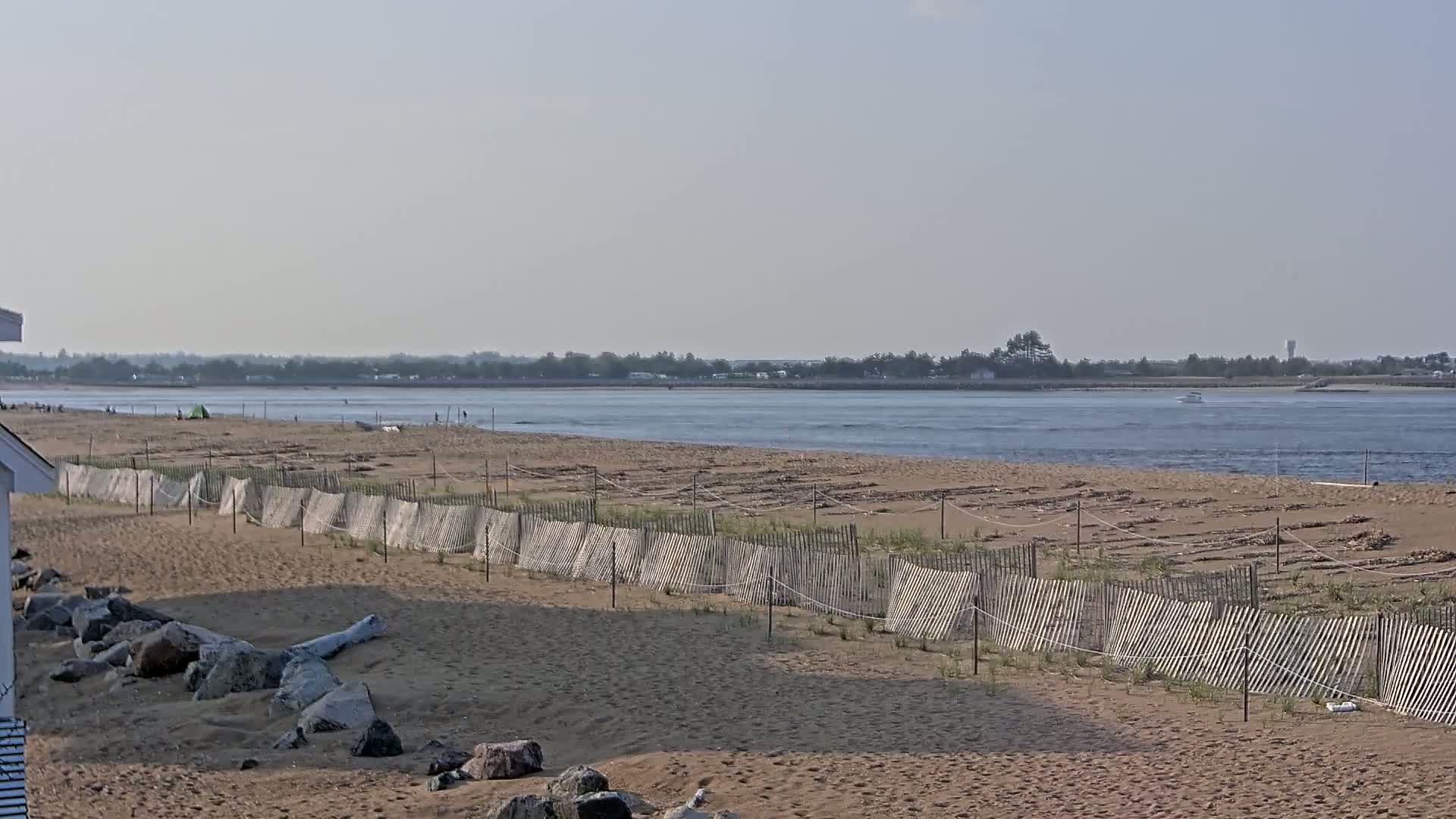 A sandy beach with a wooden fence stretches along a calm body of water under a hazy sky, with a few people visible in the distance.