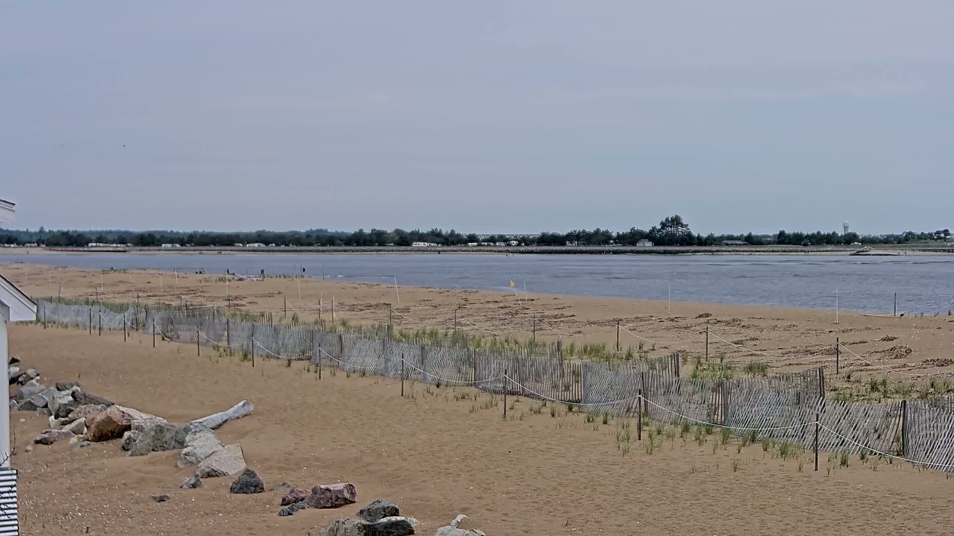 A sandy beach with a low wooden fence stretches toward a calm body of water under an overcast sky.