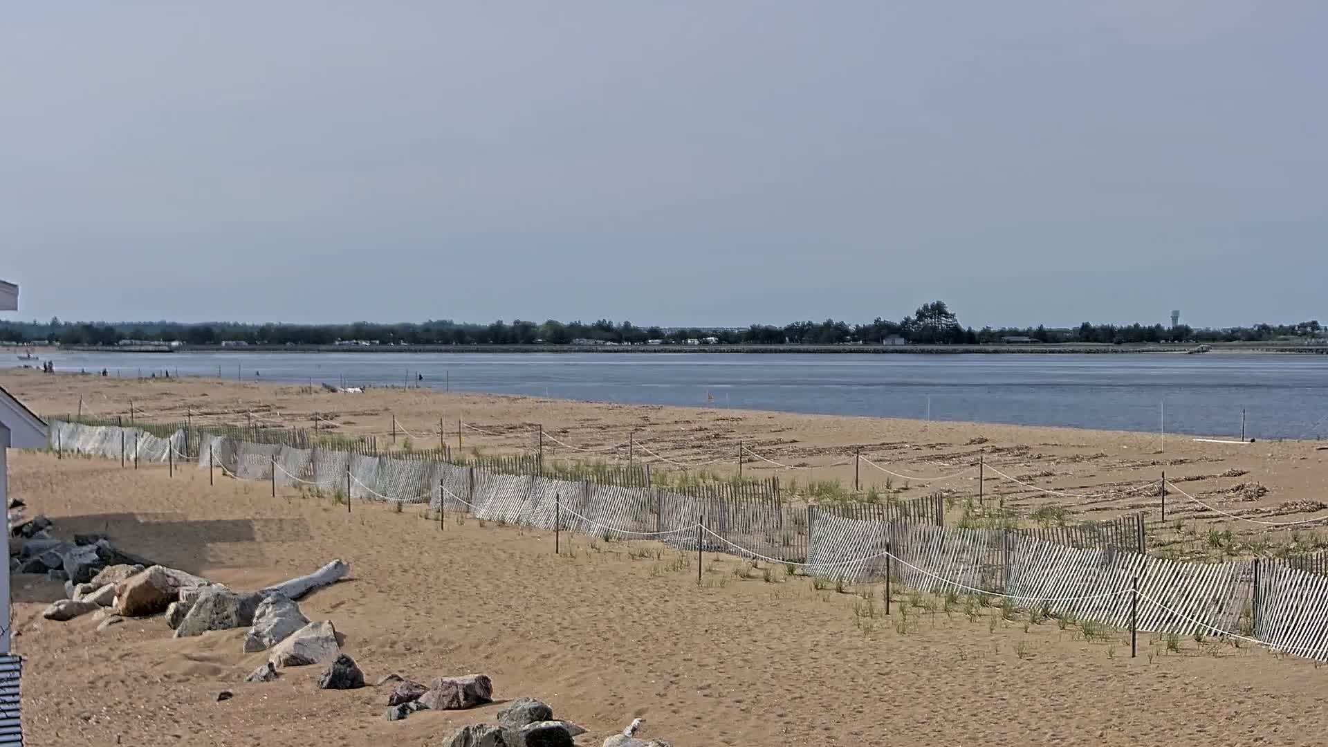 A sandy beach with a low wooden fence stretches along a calm body of water under a mostly clear sky.