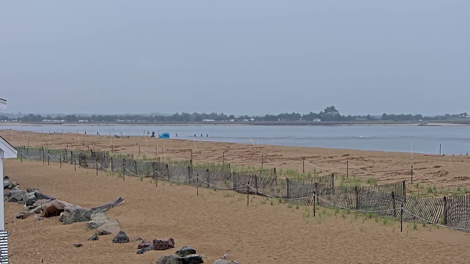 A sandy beach with a low wooden fence stretches to a calm, grey body of water under an overcast sky.