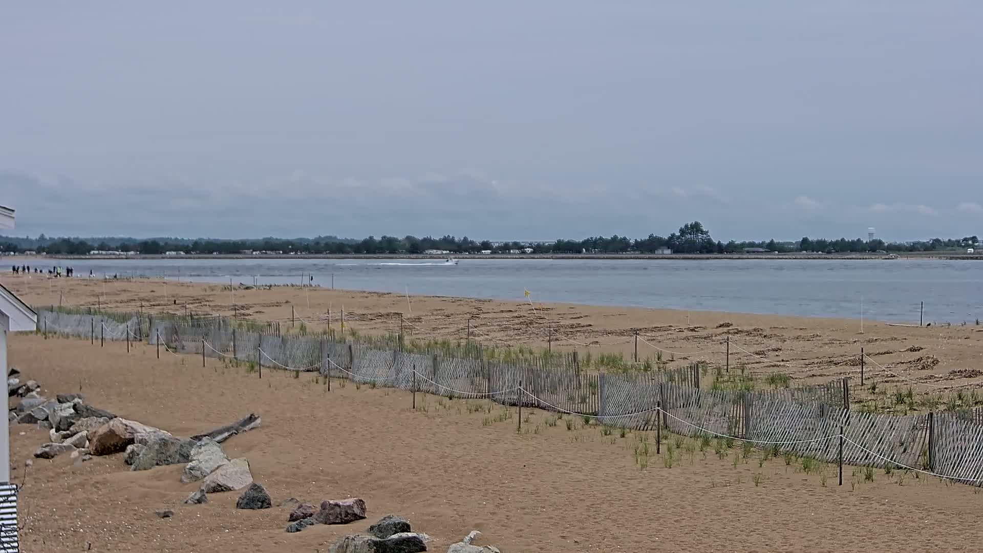 A sandy beach with a low wooden fence protecting young vegetation stretches toward a calm body of water under an overcast sky.
