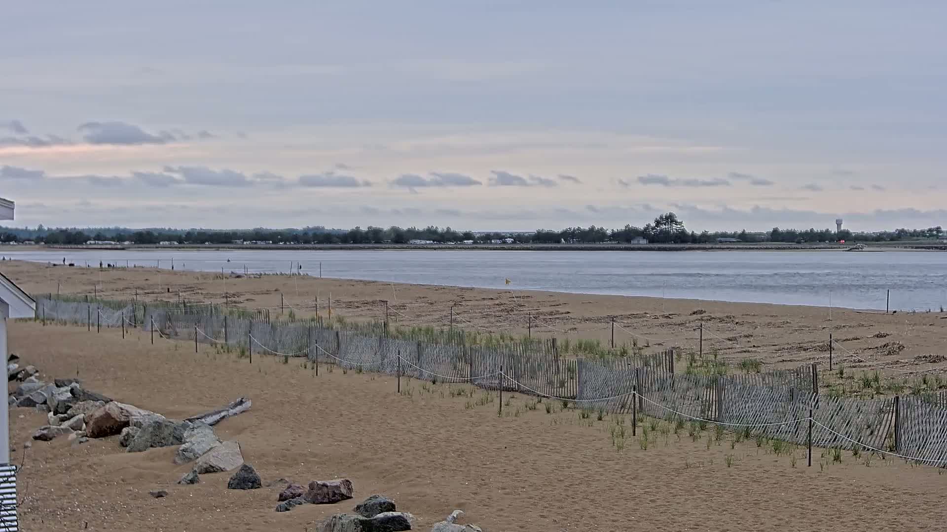A sandy beach with a low wooden fence protecting young vegetation stretches toward a calm body of water under an overcast sky.