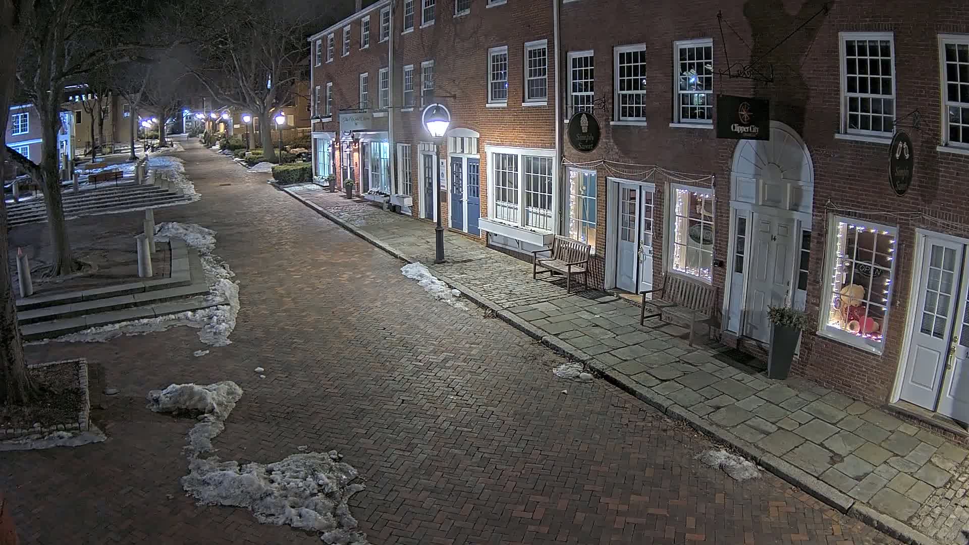 A brick street lined with brick buildings at night, with patches of snow on the ground and sidewalks.