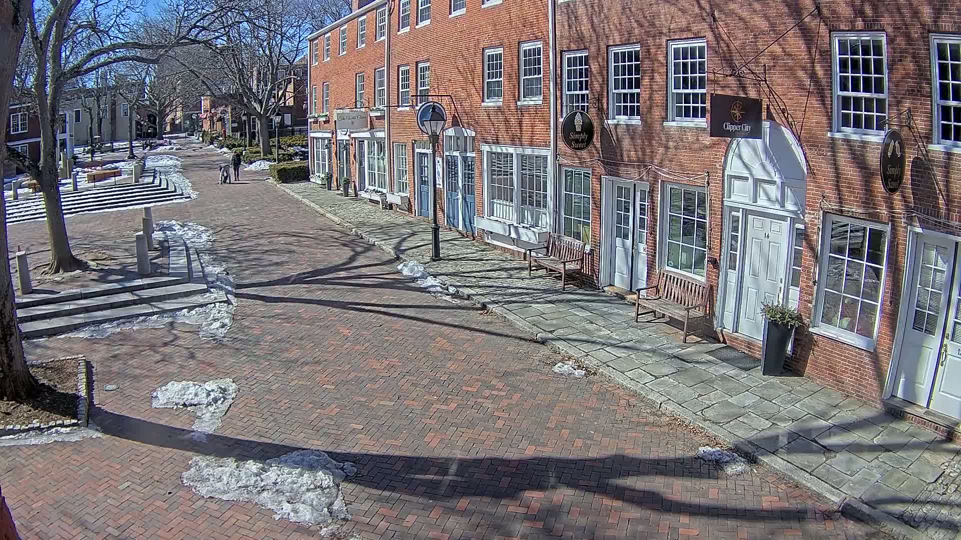 A brick-paved street lined with brick buildings on a sunny day with patches of snow and ice on the ground.