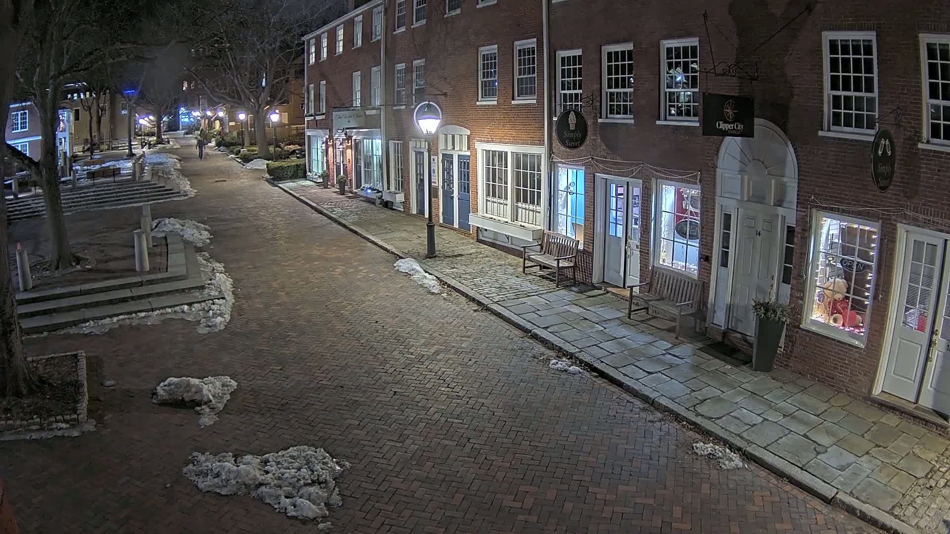A snow-dusted brick street at night lined with brick buildings, lit by street lamps.