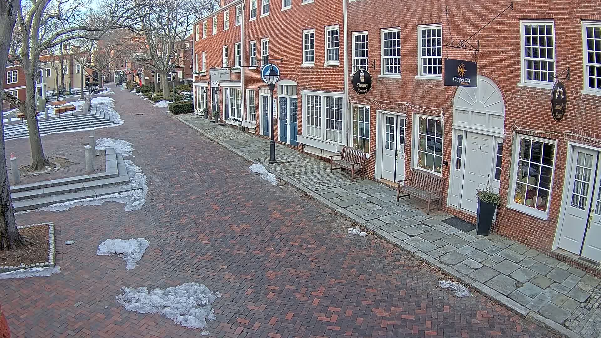 A brick street lined with brick buildings, some with storefronts, is partially covered in snow.