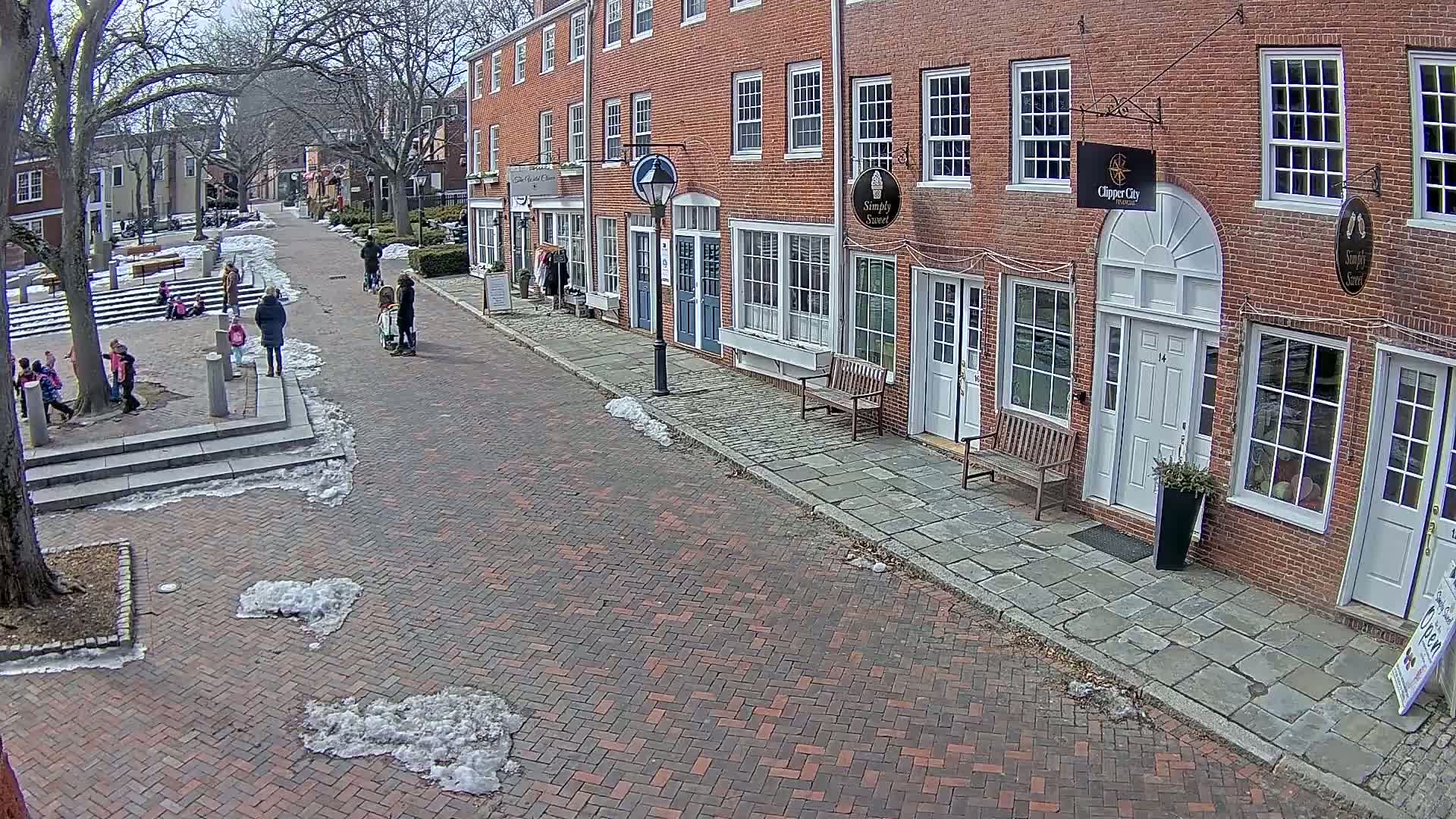 A brick street lined with brick buildings and a few people, with patches of snow and ice on the ground under a mostly clear sky.