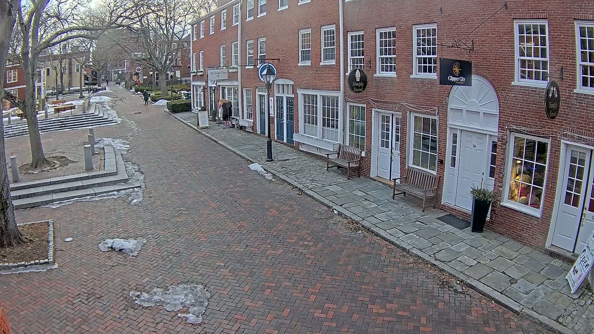 A brick-paved street lined with brick buildings, with patches of snow and ice on the ground, under a mostly clear sky.