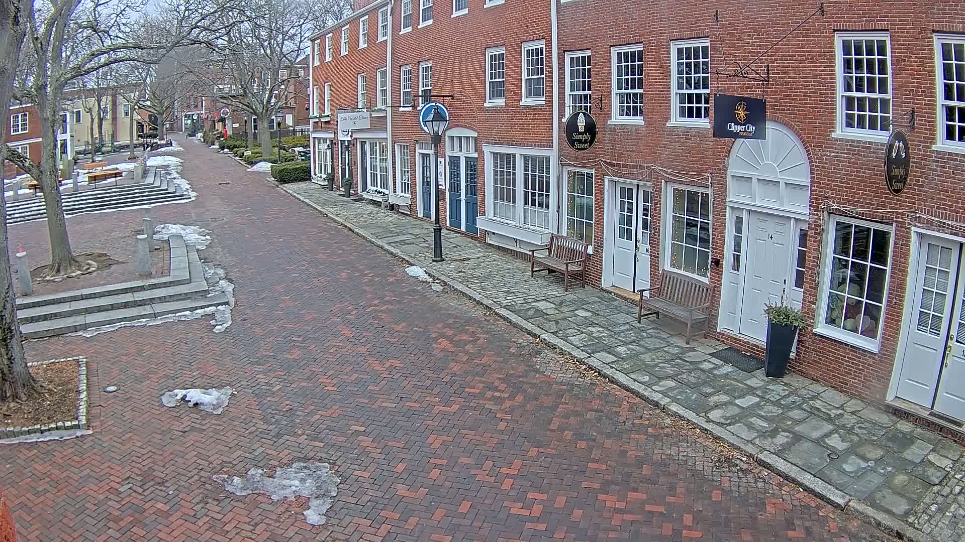 A brick street with snow patches leads to a row of brick buildings on a cloudy day.