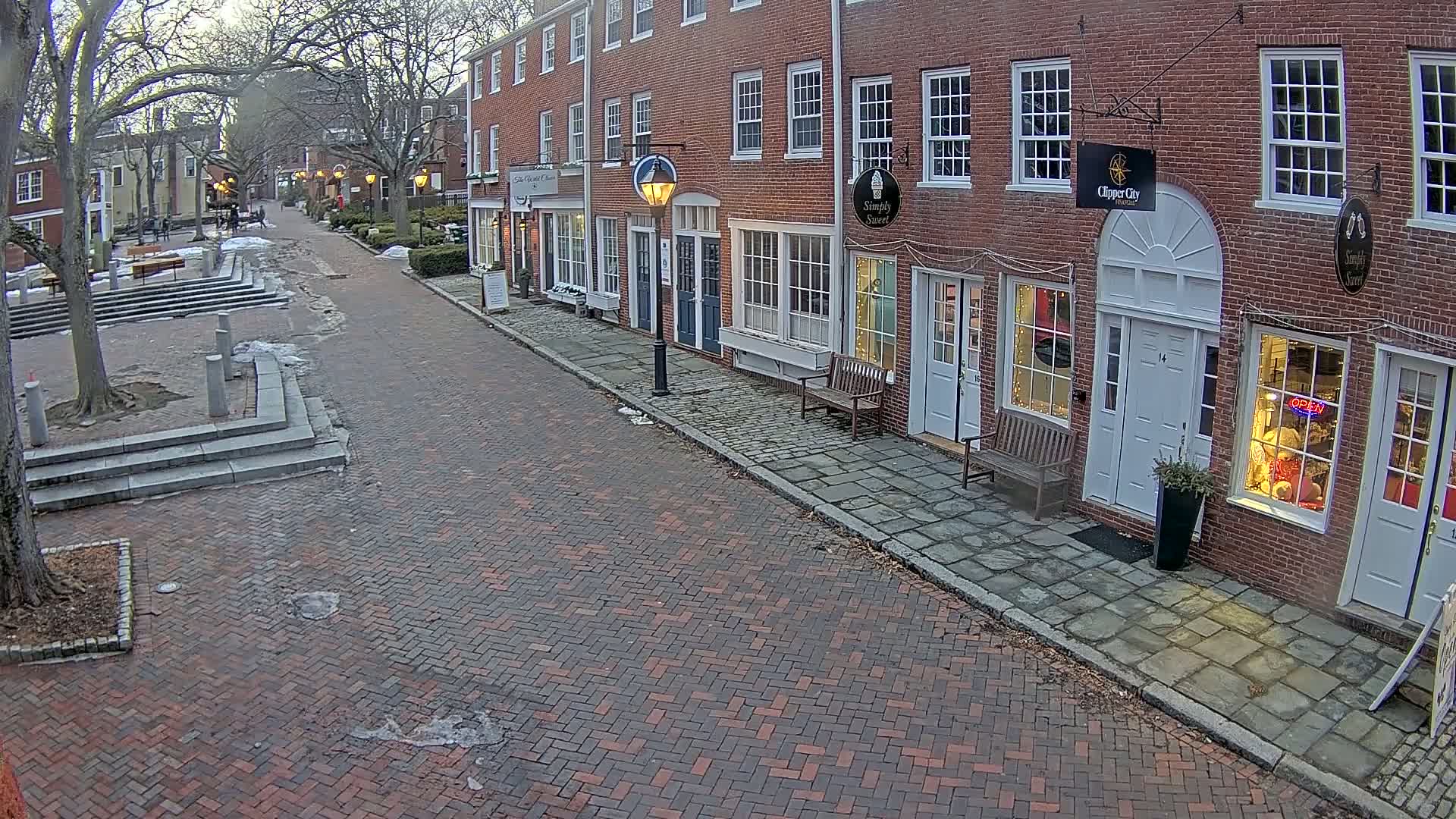 A brick-paved street lined with brick buildings on a partly cloudy, cool day, with patches of snow and ice on the ground.