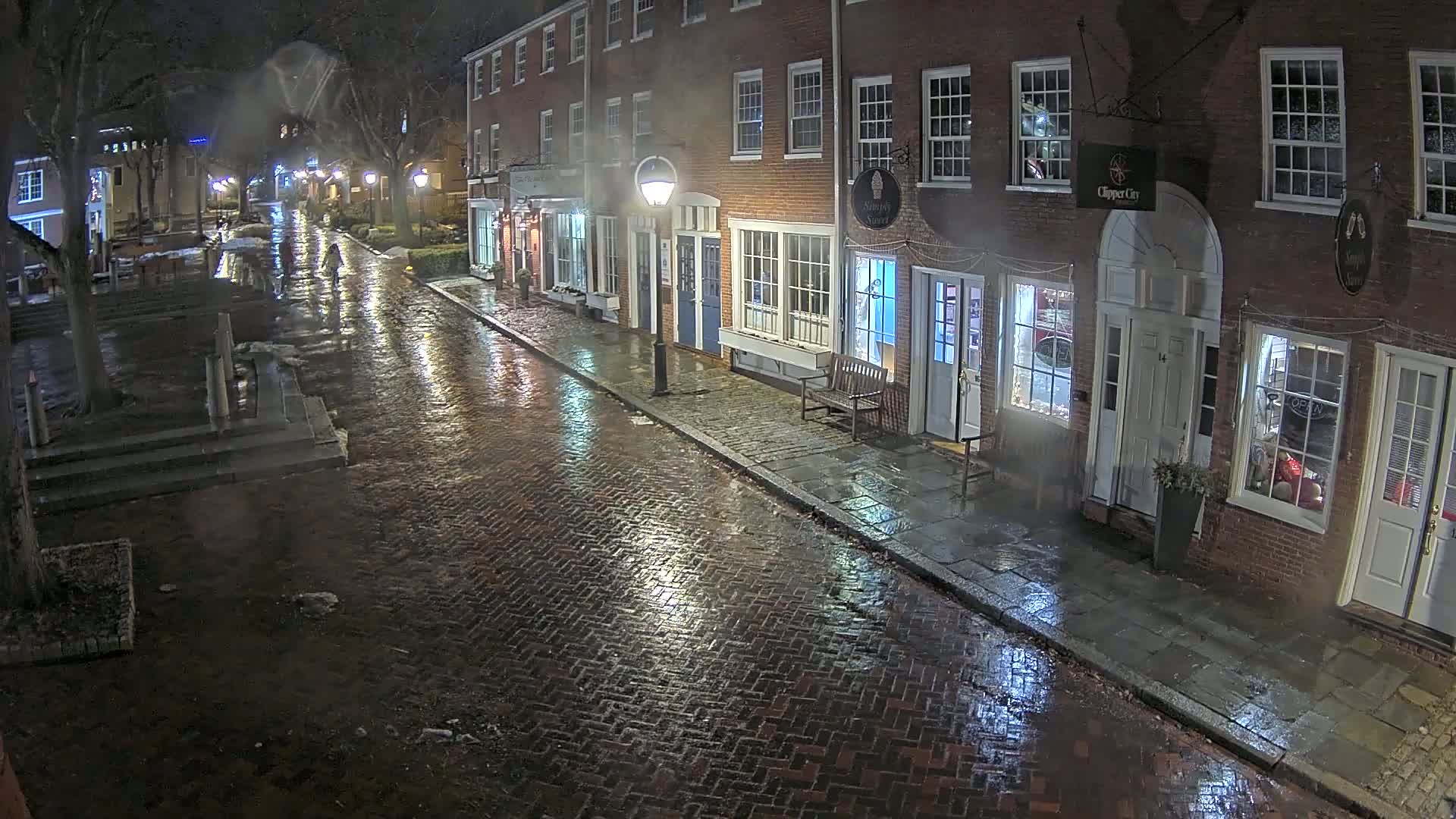A rain-slicked brick street at night is lined with illuminated storefronts in a row of brick buildings.
