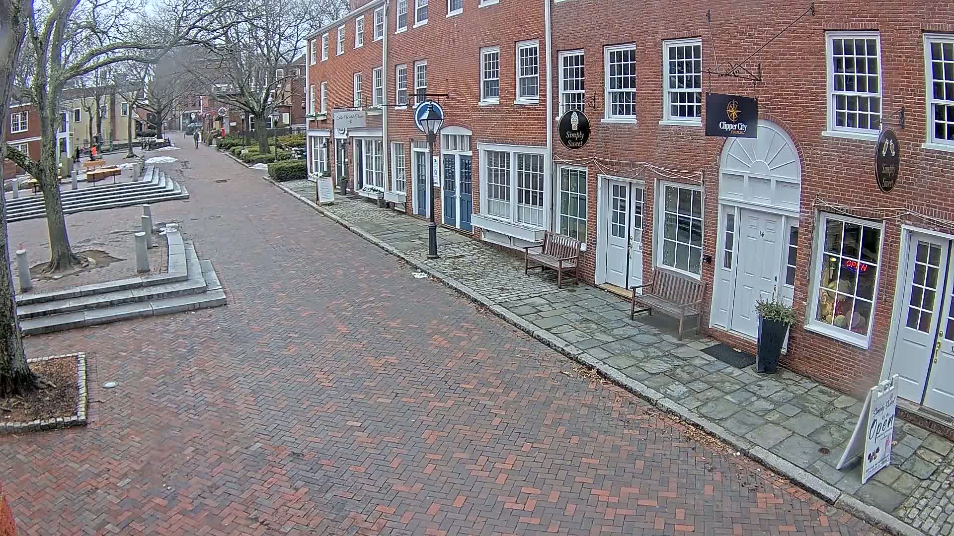 A brick-paved street lined with brick buildings on a cloudy day leads to a small town square with stone steps.