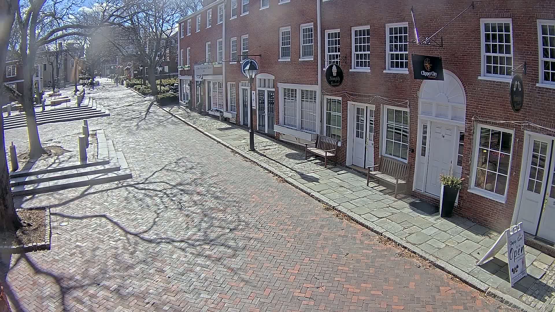 A brick street lined with brick buildings on a sunny day, with the shadows of bare trees cast across the street and sidewalks.