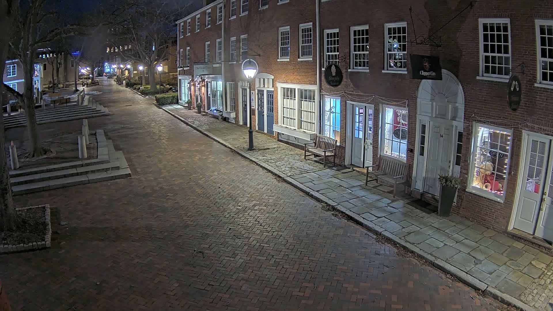 A brick-paved street at night, lined with illuminated brick buildings, is empty of people except for one visible in a window.
