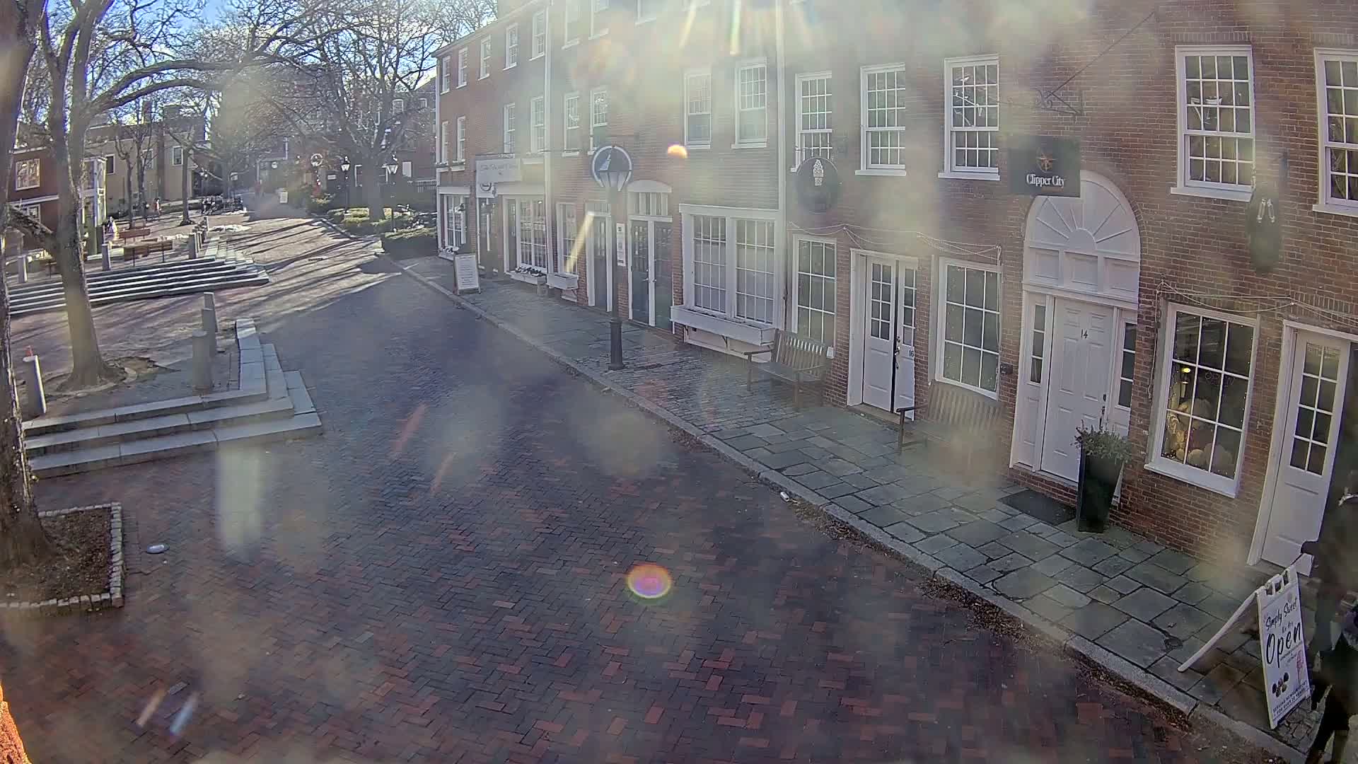 A sunny day reveals a brick street lined with brick buildings, some with shopfronts, and a small plaza with trees and steps in the background.