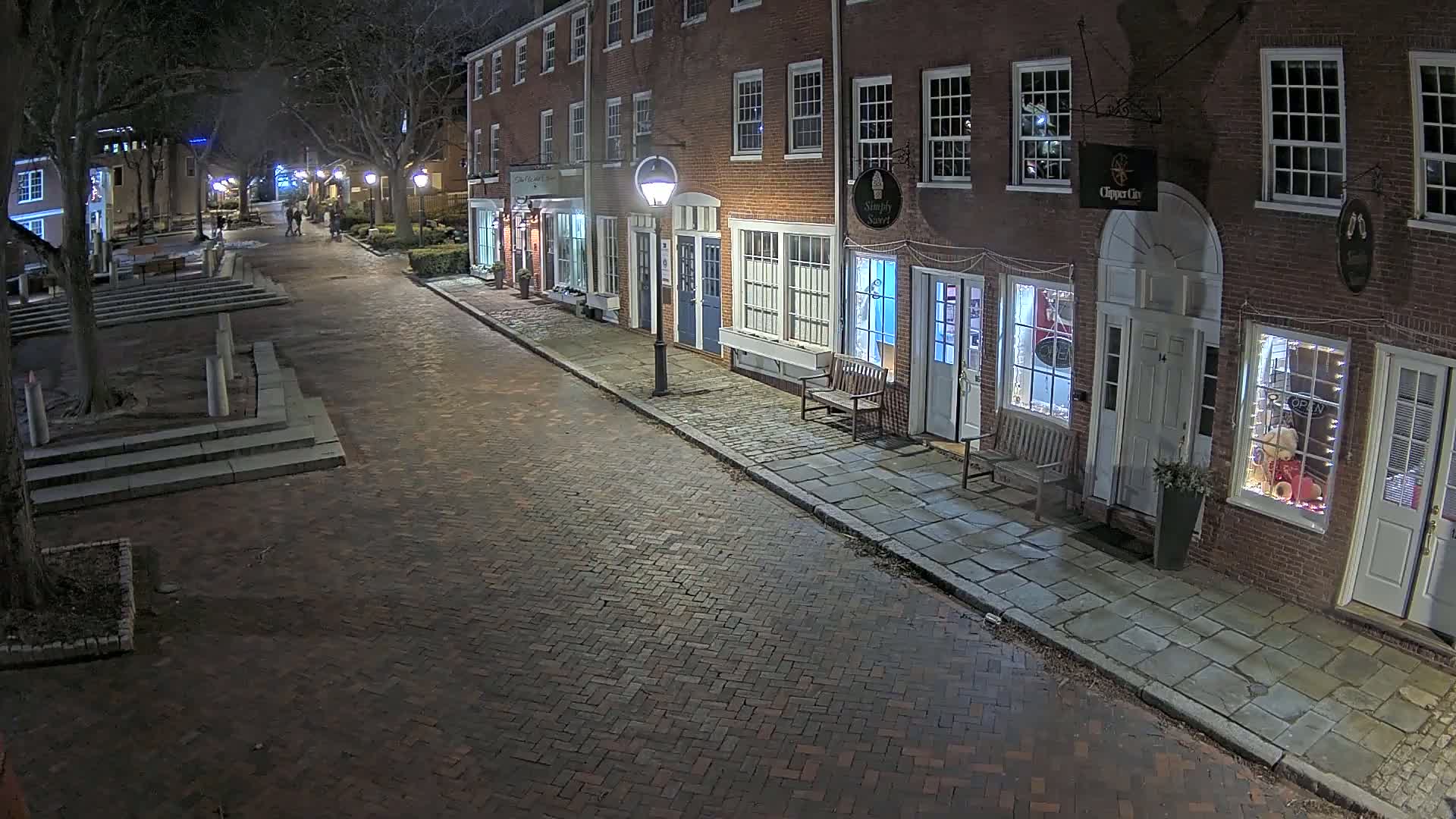 A brick street at night lined with brick buildings, some with illuminated shop windows, is sparsely populated with pedestrians.
