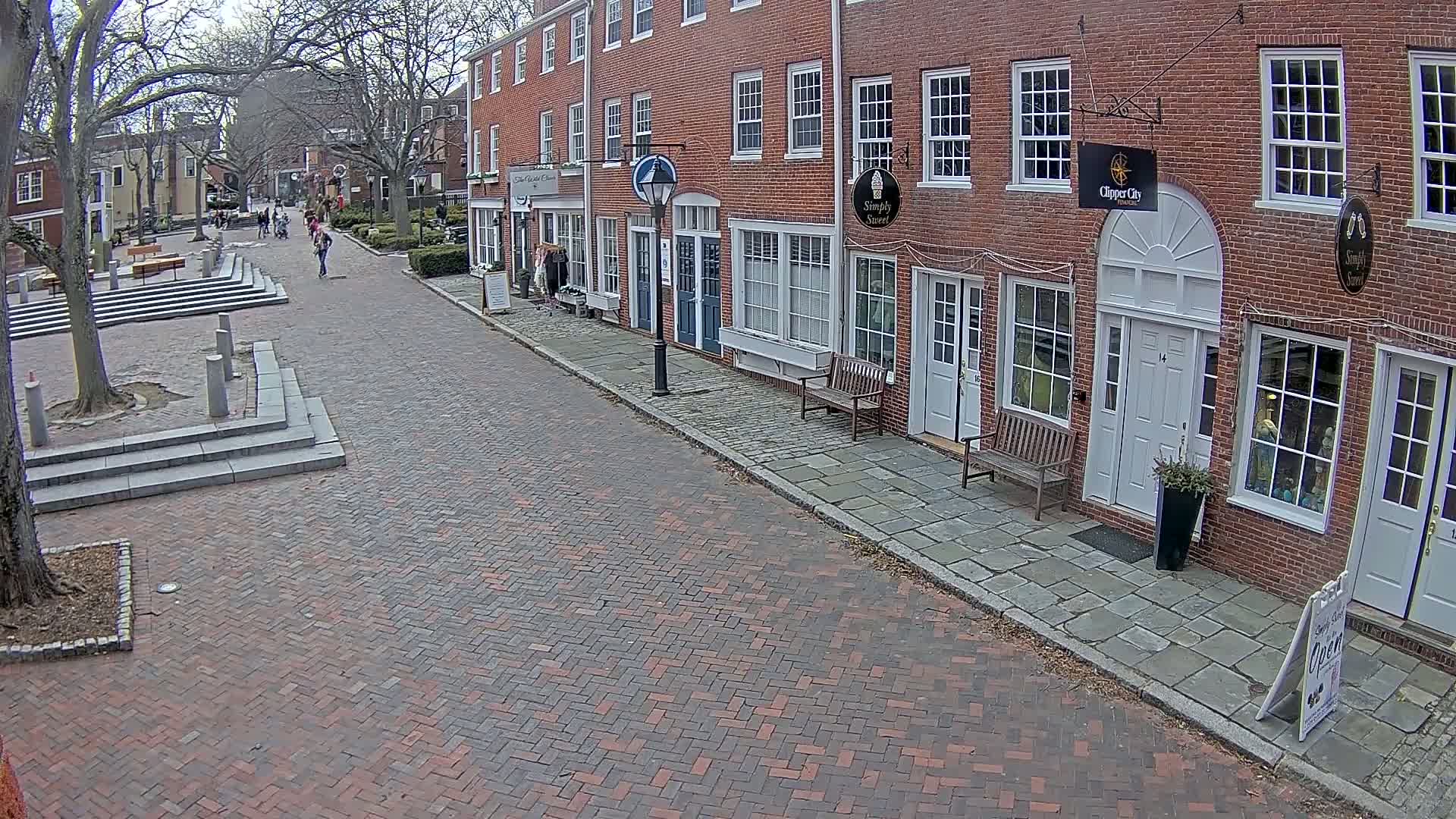 A brick-paved street lined with brick buildings under a mostly cloudy sky, with a few pedestrians visible in the distance.