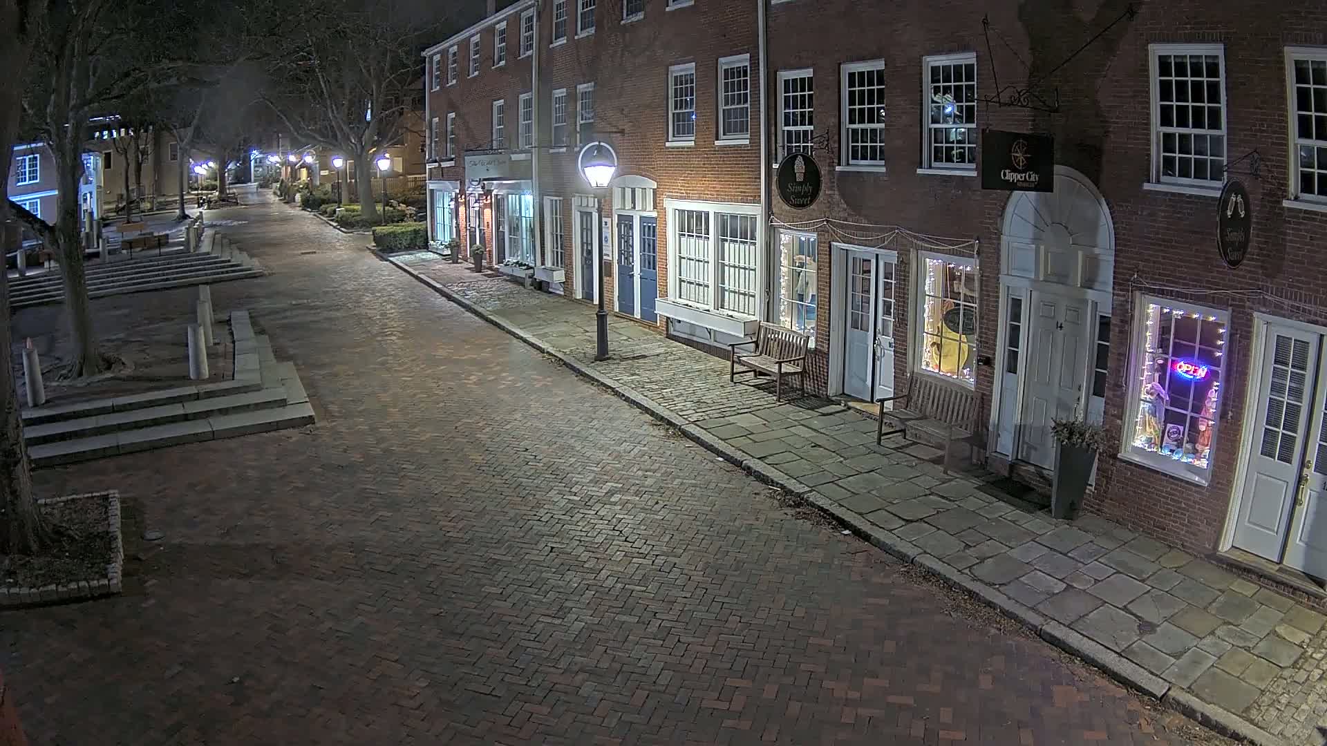 A brick-paved street at night, lined with illuminated storefronts in a row of brick buildings, is sparsely populated under the cover of darkness.