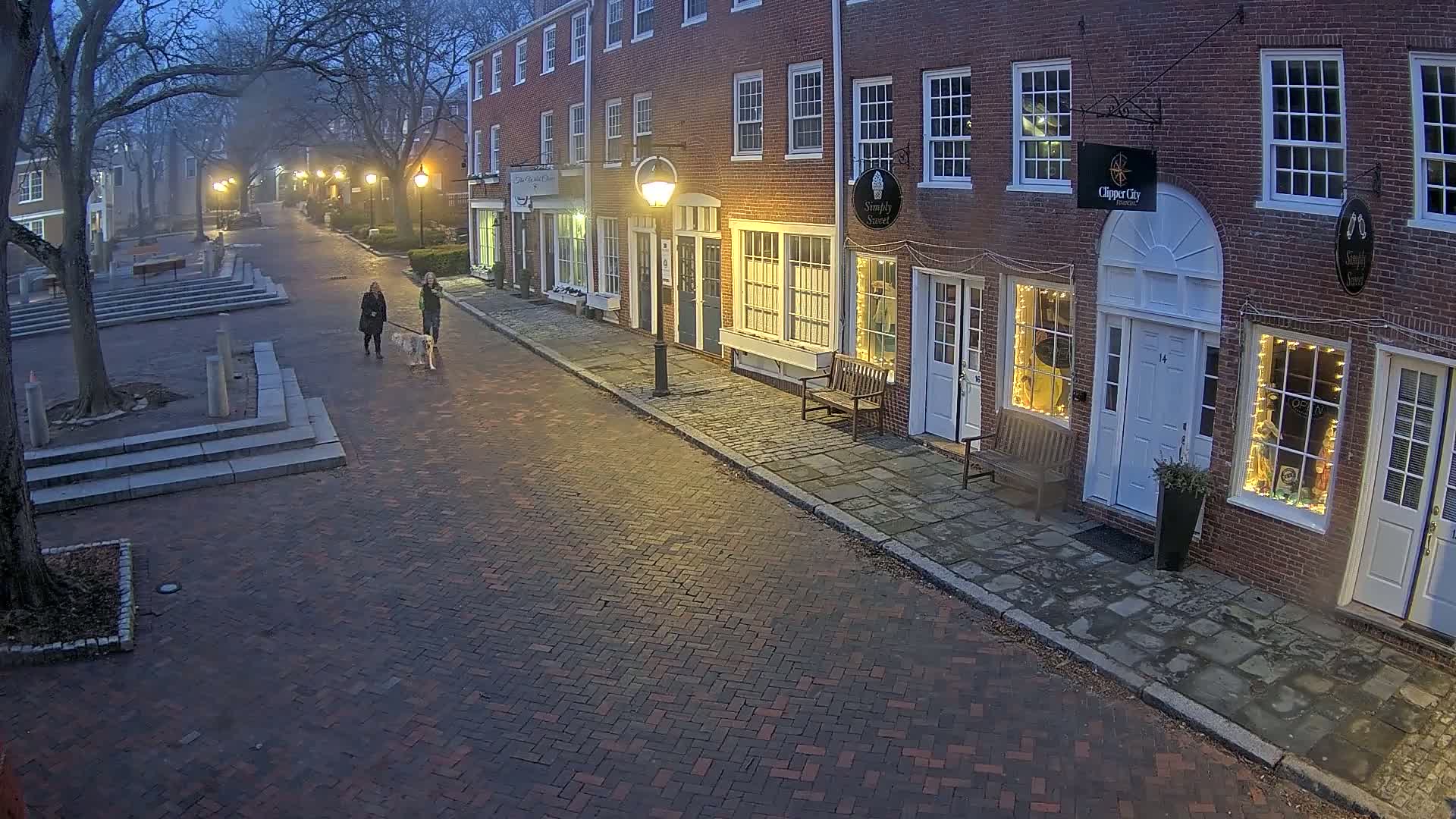 Two people walk a dog down a brick-paved street lined with brick buildings at dusk under dim lighting.