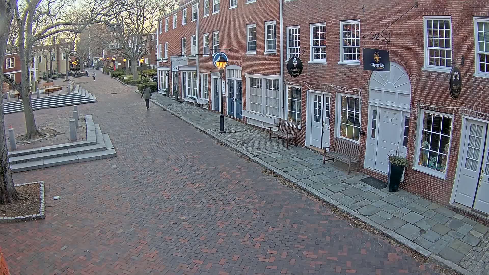 A brick-paved street lined with brick buildings under a mostly cloudy sky, with a single pedestrian walking away.