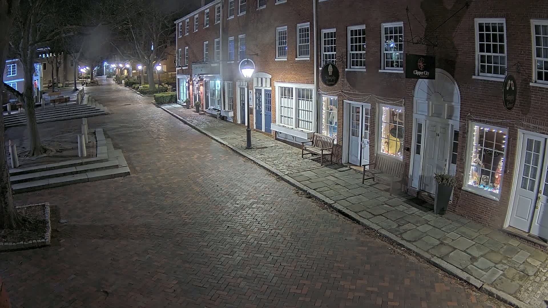 A brick street at night, lined with illuminated storefronts in a row of brick buildings, under a slightly hazy sky.