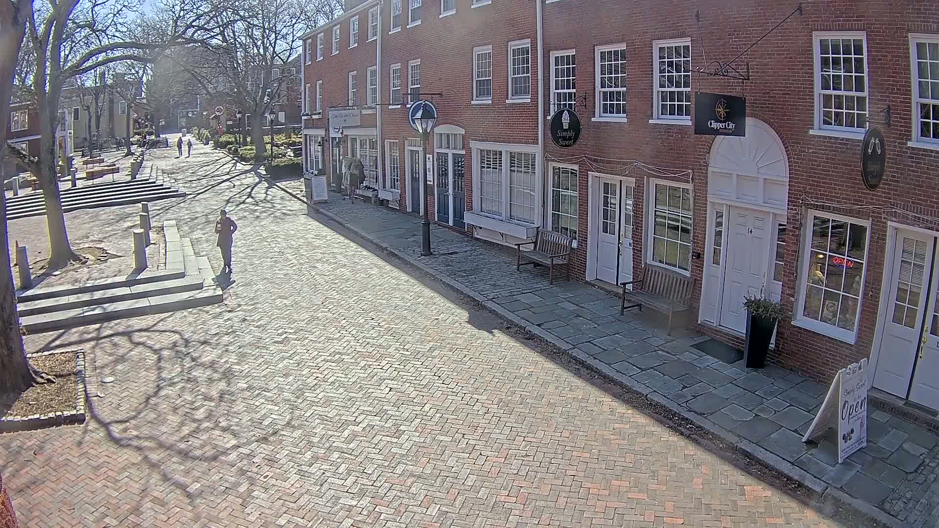 A brick-paved street lined with brick buildings on a sunny day, with a few pedestrians visible in the distance.