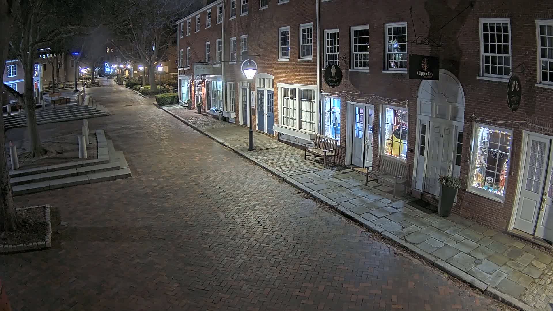 A brick-paved street at night, lined with brick buildings containing shops, is lit by street lamps under a clear, dark sky.