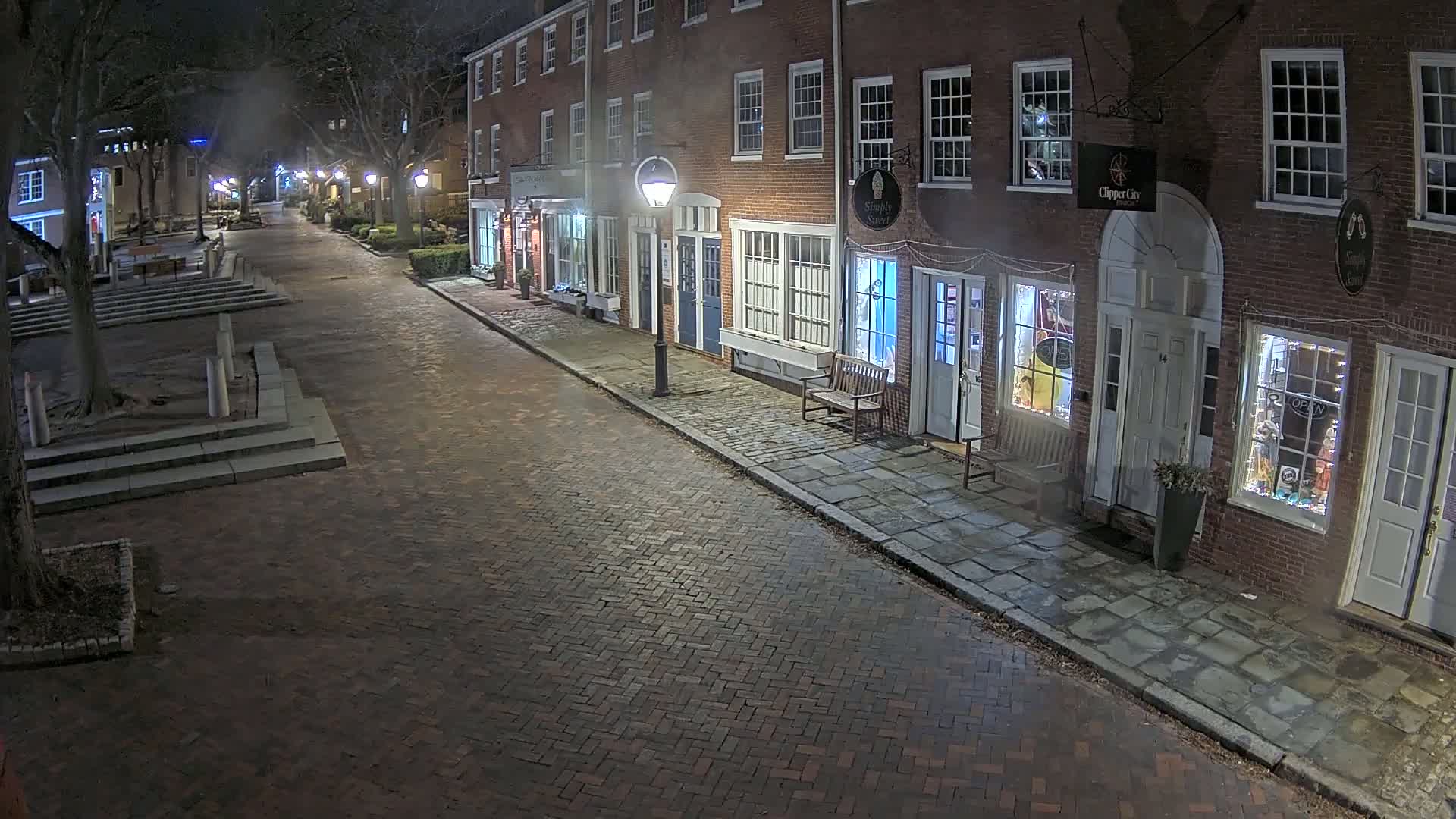 A brick street at night, lined with illuminated storefronts in a row of brick buildings, is mostly empty except for a park-like area with stairs to the left.
