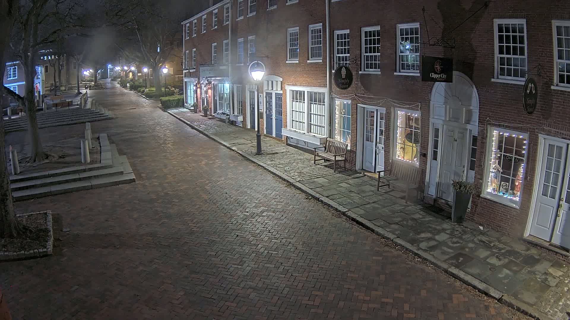 A brick street at night, lined with illuminated storefronts in brick buildings, is lightly misted with fog.