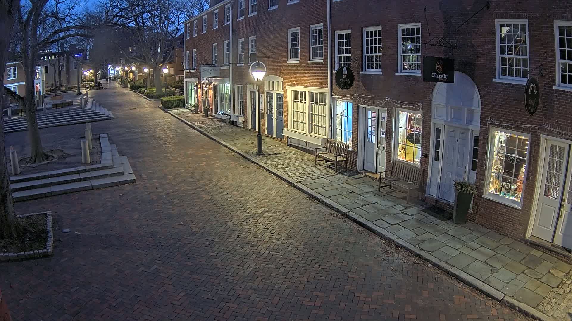 A brick-paved street at night lined with illuminated storefronts in a row of brick buildings, under a dark sky.