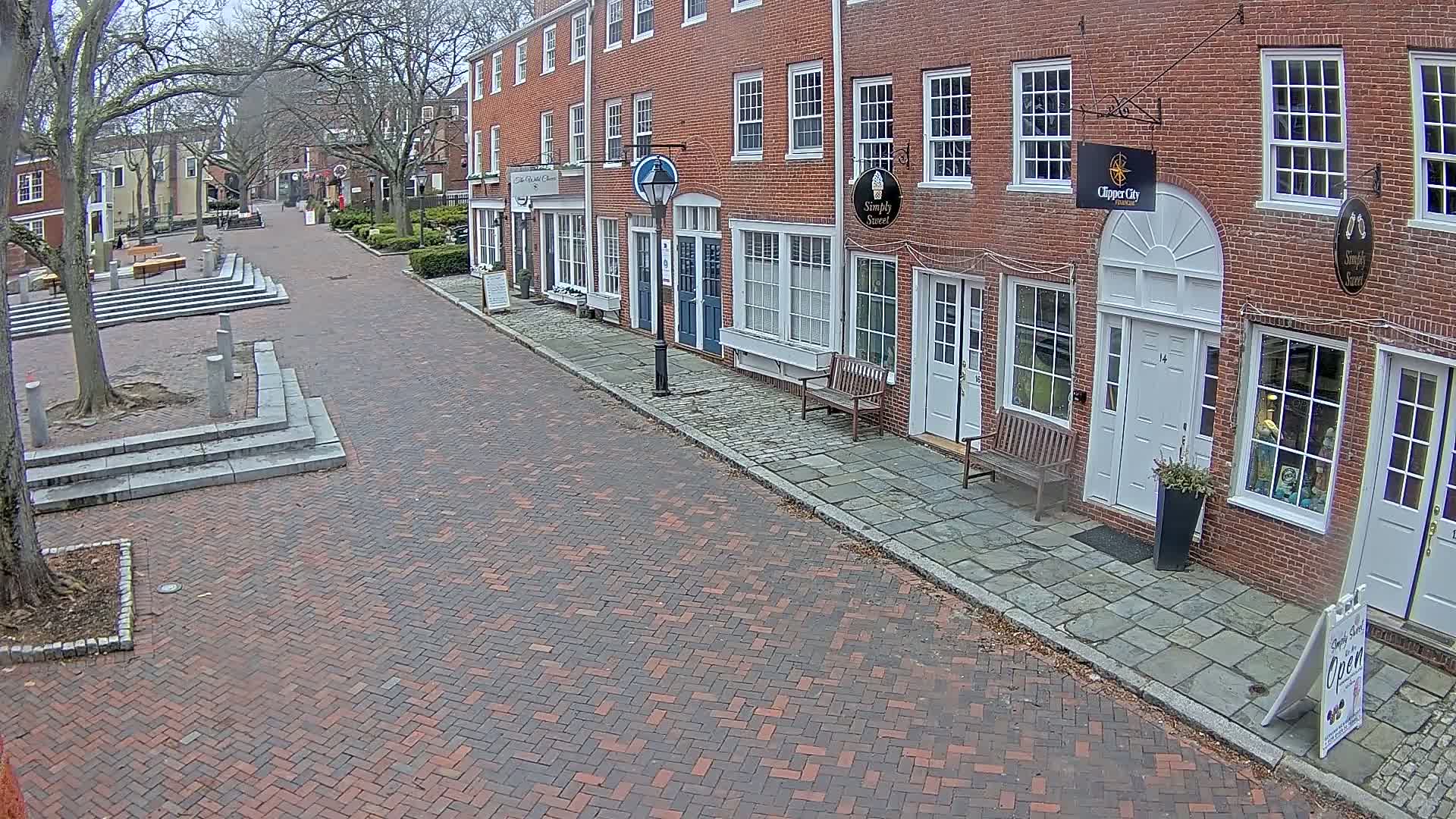 A brick street lined with brick buildings, some with benches outside, on an overcast day.