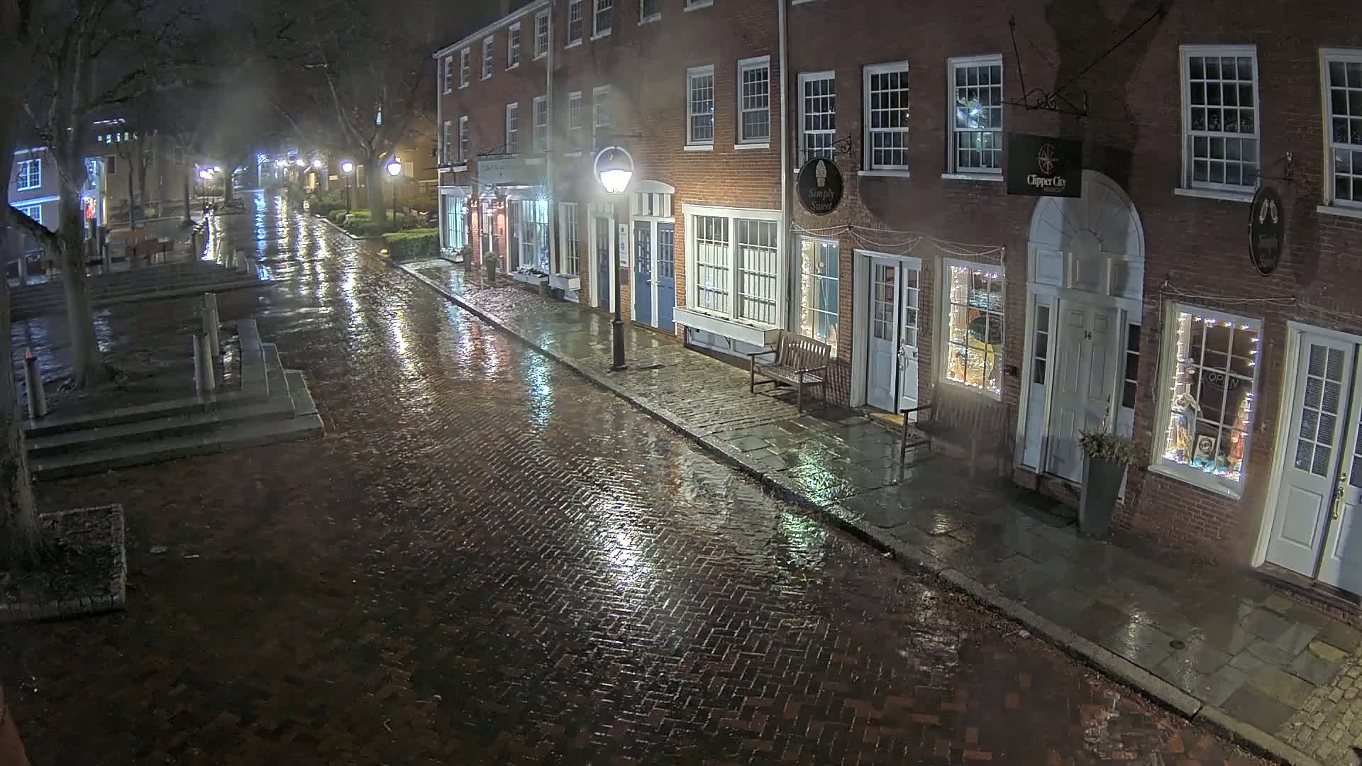 A rain-slicked brick street at night, lined with illuminated storefronts in a row of brick buildings.