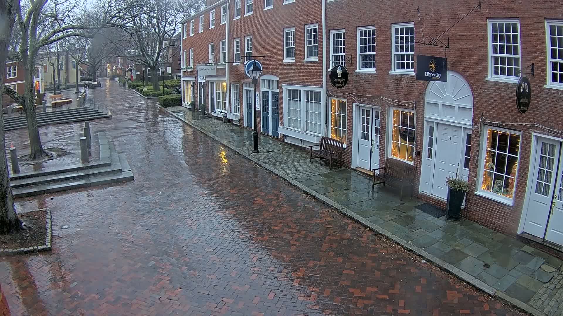 A rain-slicked brick street leads past several brick buildings with shopfronts, under a gray sky.