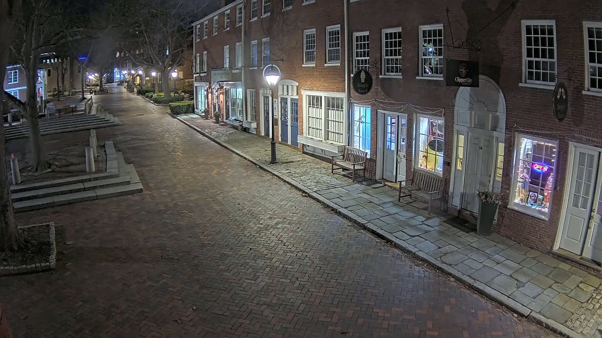 A brick street at night, lined with illuminated storefronts in brick buildings, and bordered by trees.
