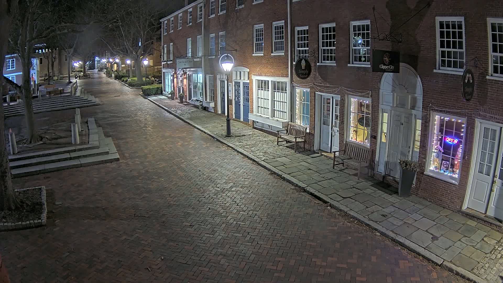 A brick street at night lined with brick buildings, some with lit windows and a few streetlights illuminating the scene.